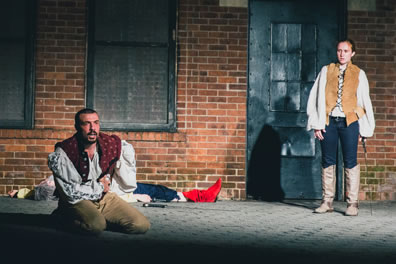 Hotspur kneeling on the ground, hands clutching his side, wearing red vest over white puffy shirt and tan pants, Hal standing off to the right, sword in her left hand, wearing buckskin vest over white puffy shirt, black jeans and gray knee-high boots, Falstaff lying in the background (red boots), sword next to him, all on a brick surface, and in the background the Carroll Park utility building, brick with black doors and windows.